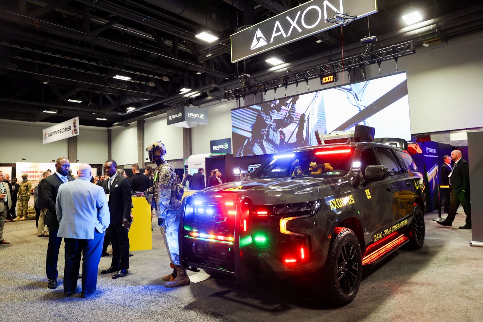 An Axon vehicle with its lights on beside a soldier mannequin, attendees talking nearby